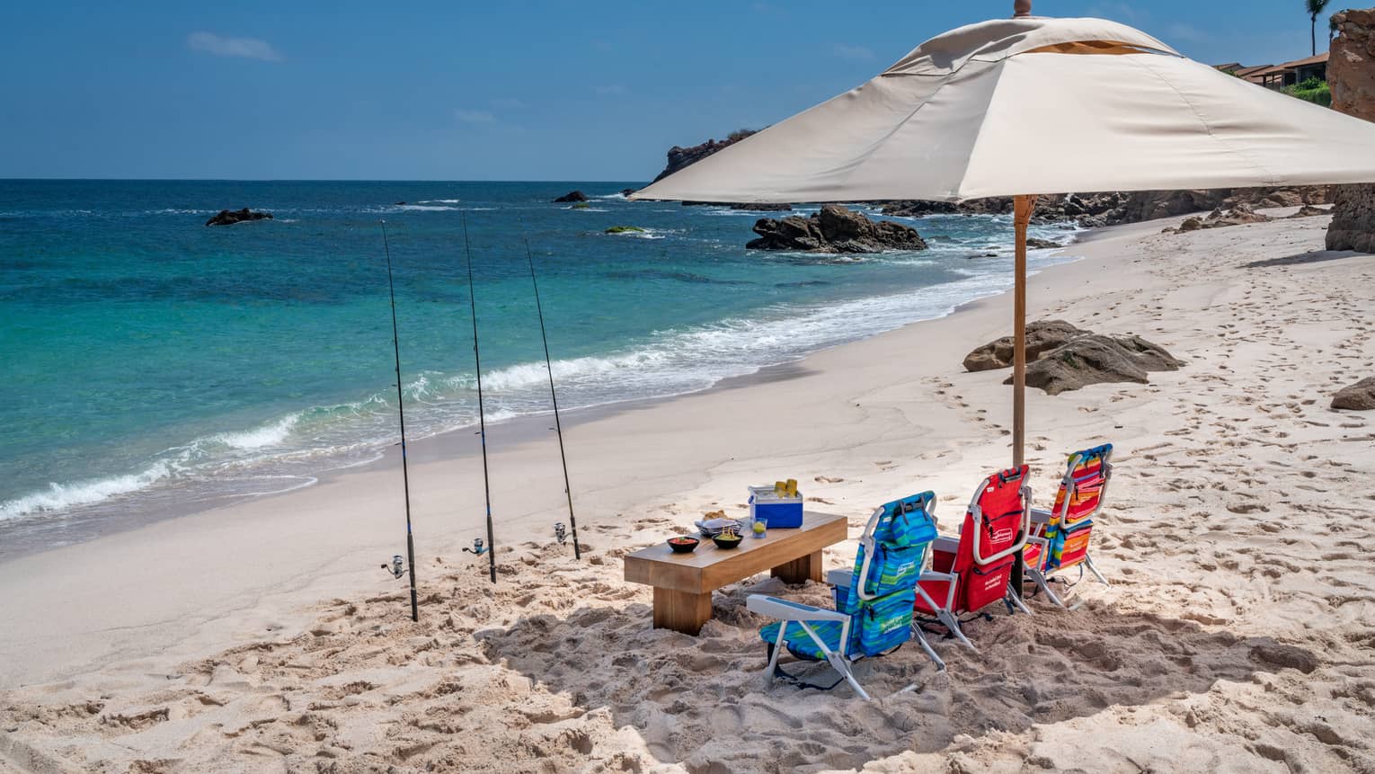 Under an umbrella, drinks, snacks and three colourful beach chairs. Three fishing rods planted in the sand, blue sea ahead.