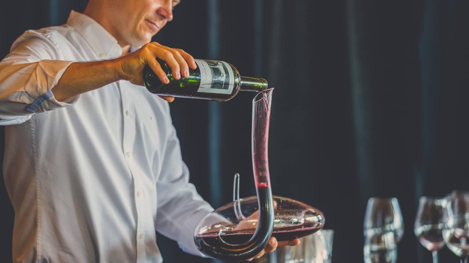 Man pours red wine into long, curved glass carafe near wine glasses