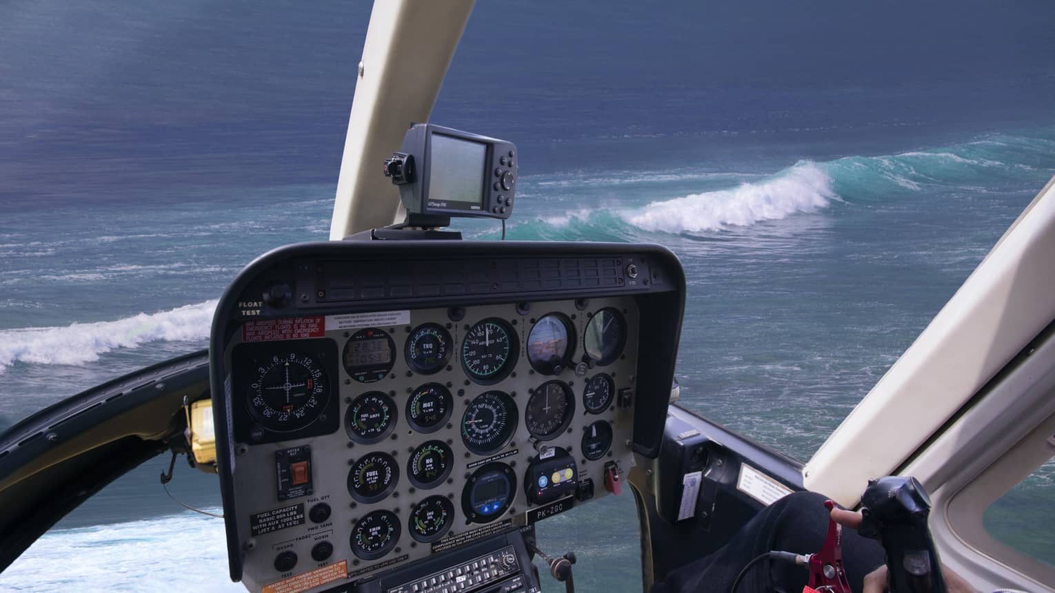 Pilot’s view of a helicopter control panel below a windshield looking out at white-capped waves in a variegated blue sea.