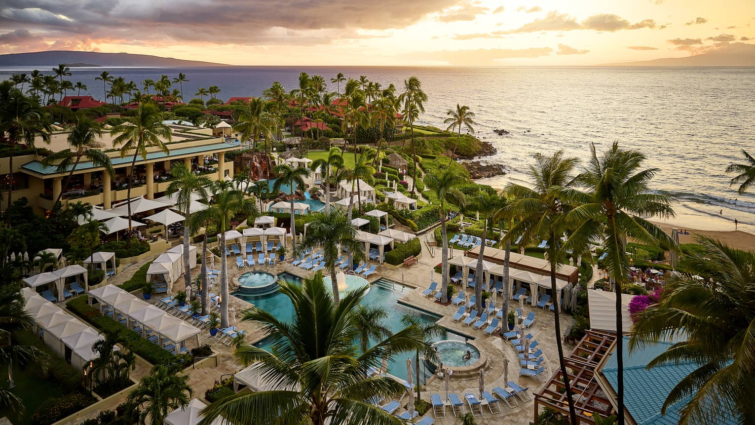 Tropical luxury resort pool lined with blue lounge chairs and palm trees, set next to ocean at sunset