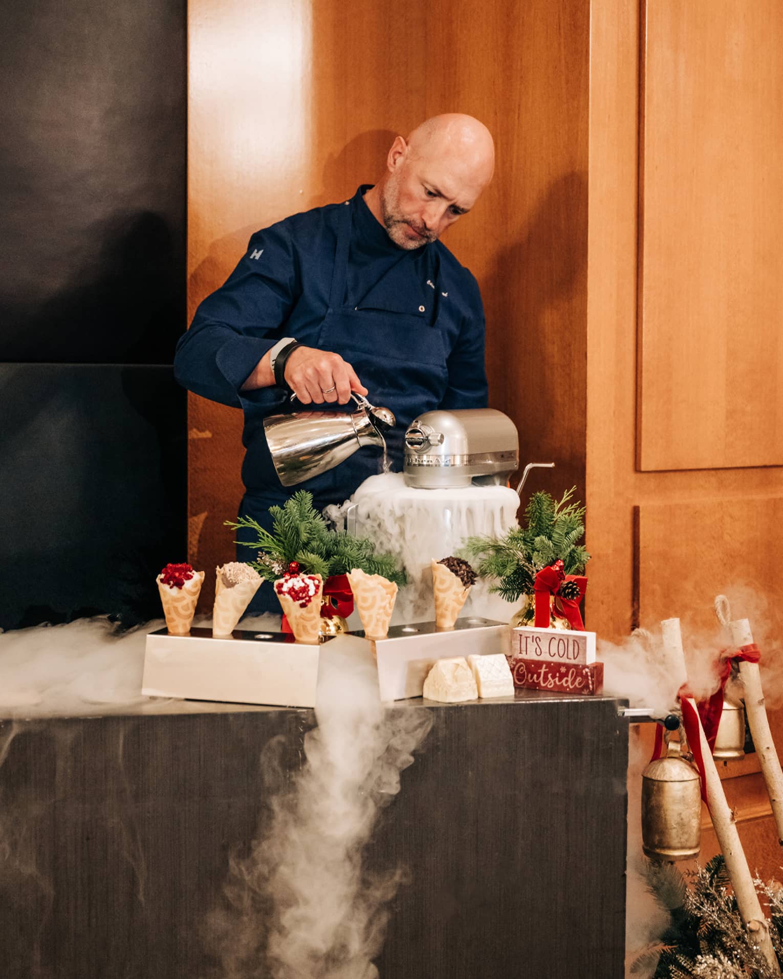 Chef pours liquid nitrogen into a mixer in front of a cart topped with waffle cones and holiday decor