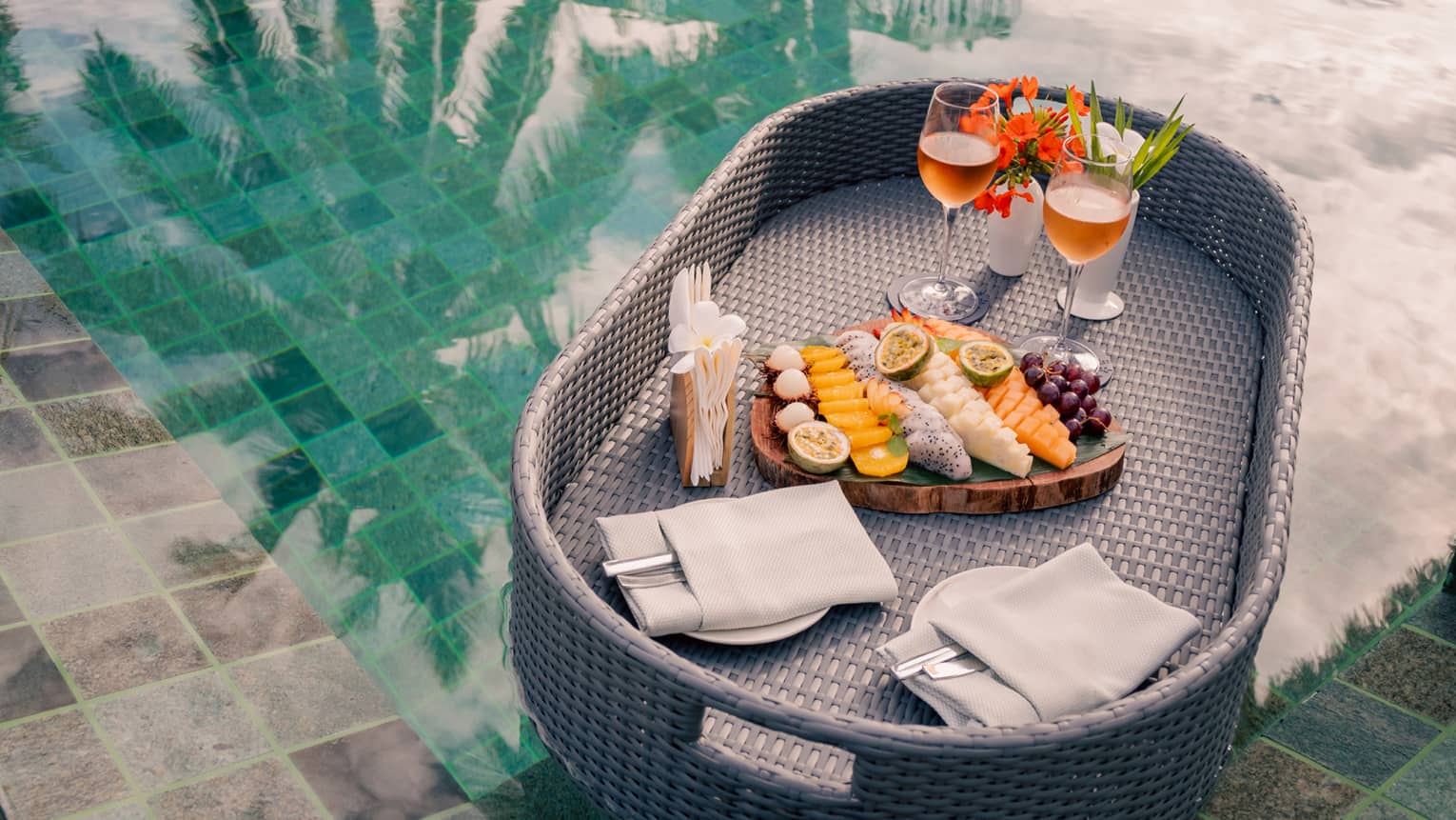 A grey wicker tray floats in a pool carrying a board of fruit arranged in rows, two plates and two glasses of rosé. 