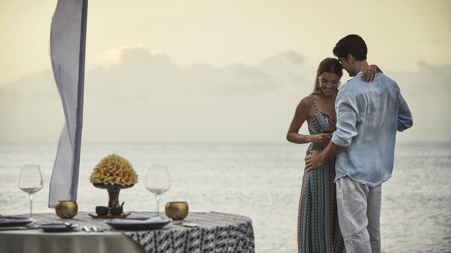 Couple embraces beside private dining table overlooking ocean at sunset