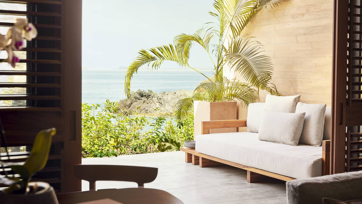 Terrace with a wooden daybed and lush potted plants, offering a view of the ocean and rocky coastline. The space is framed by open wooden shutters.
