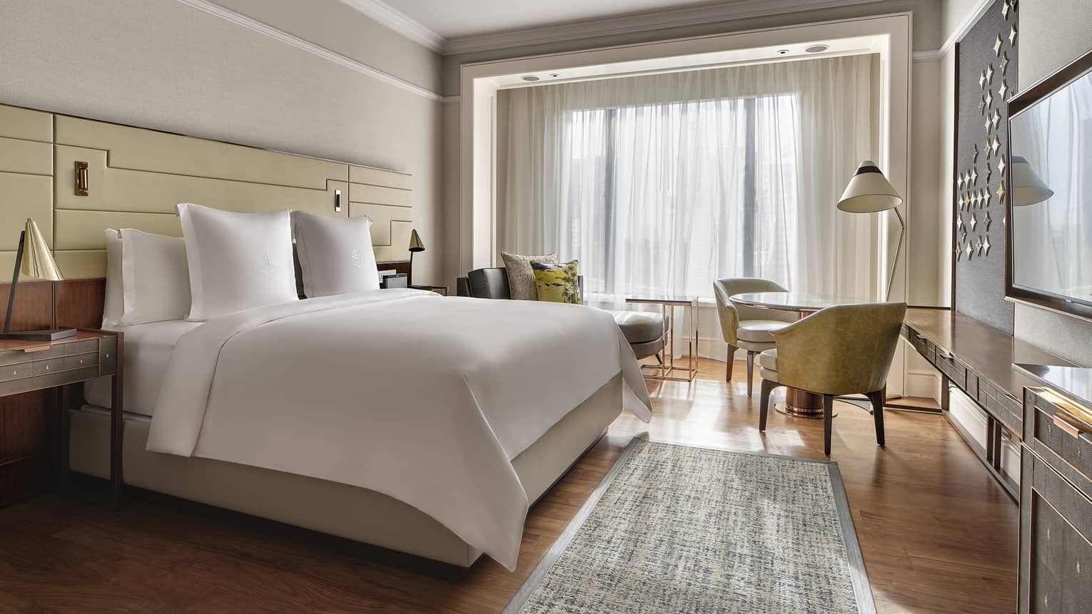 Light-filled hotel room with white king bed, wood floors and rug, two arm chairs, sheer curtains