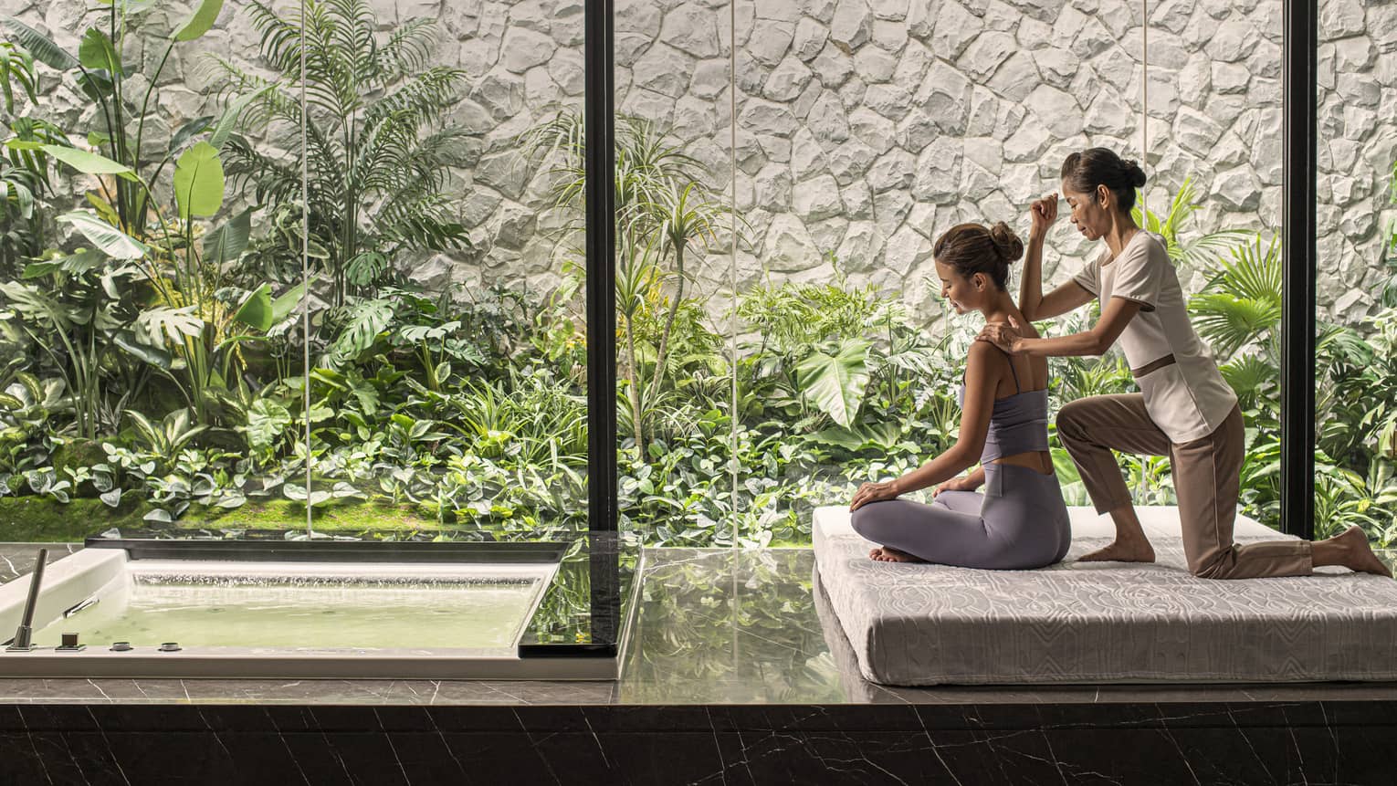 Person receiving a massage in a peaceful spa setting with large windows overlooking lush green plants and a small water feature.