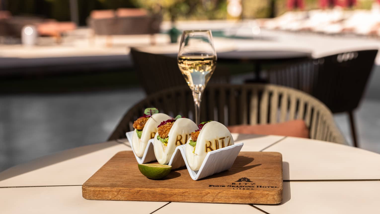 Three food items consisting of a white layer folded to hold a filling, set on a wood serving board next to a pale drink in a clear stemmed glass, all on a white table