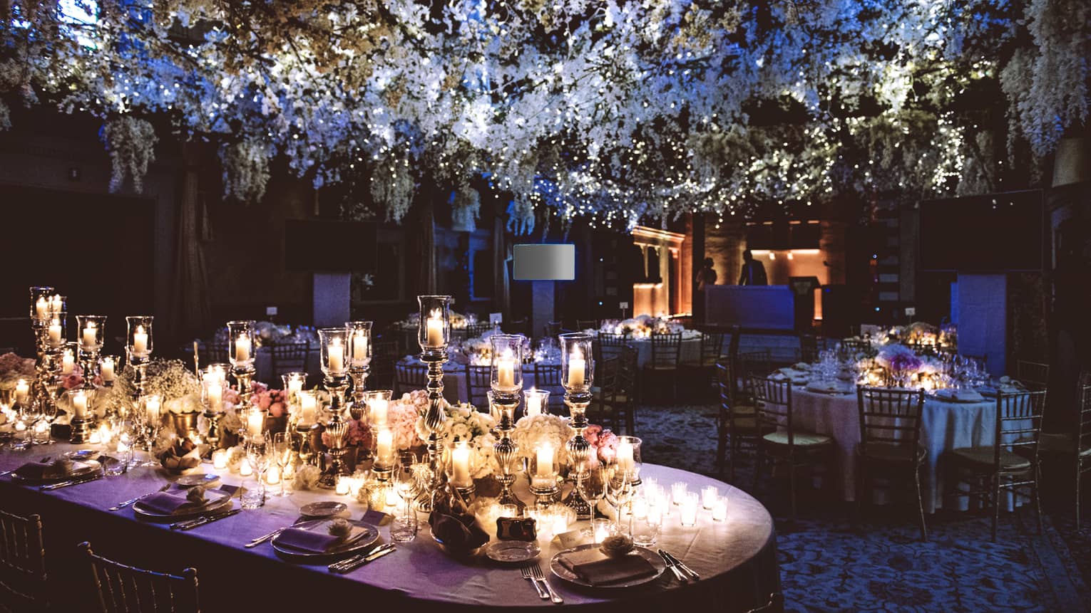 Long banquet dining table with white linens covered in glowing gold candles under flower canopy