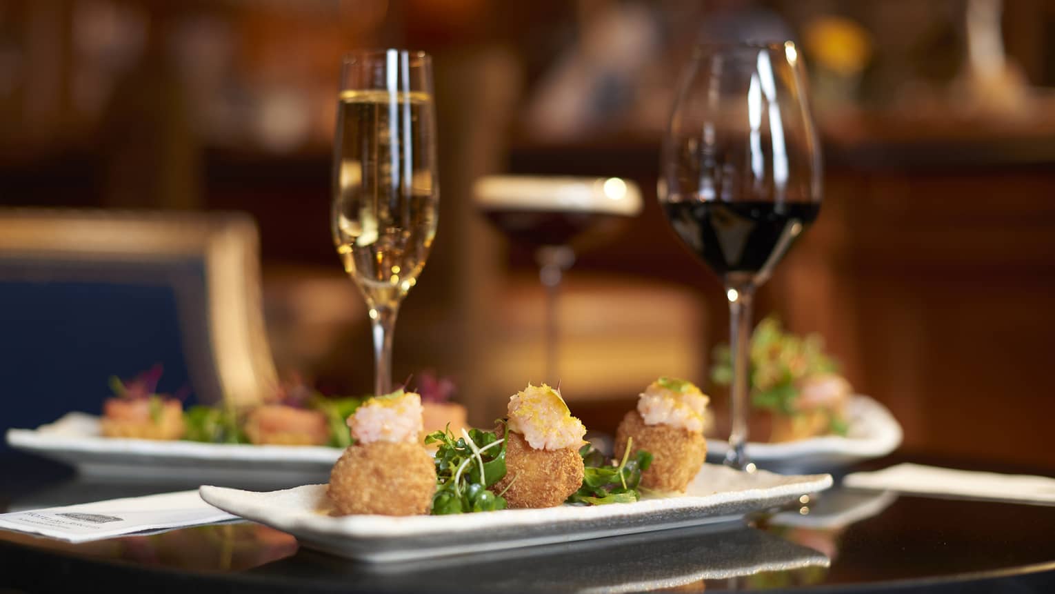 Three arancini on a white rectangular plate on a restaurant table with two glasses of wine