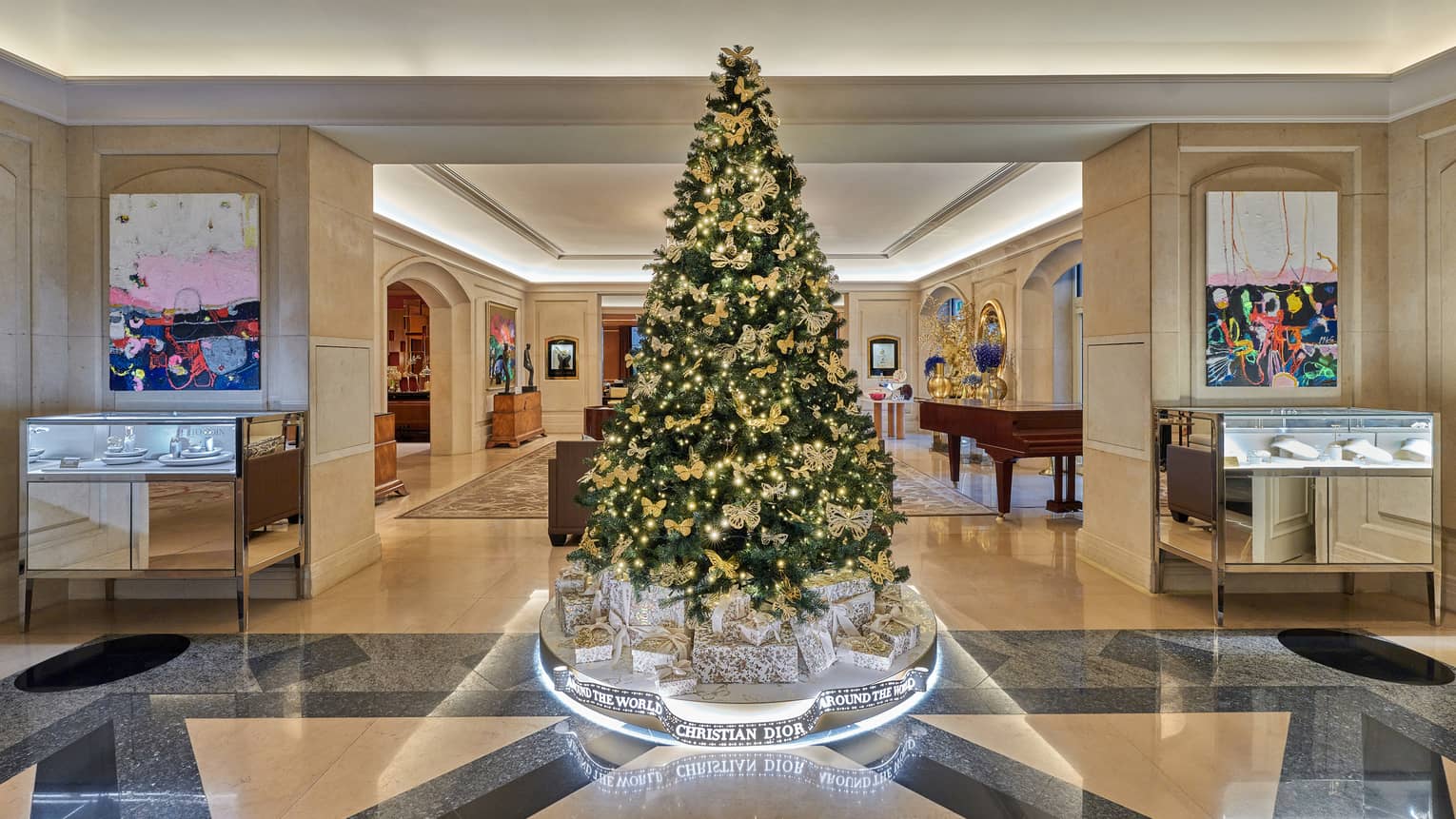 Luxurious hotel lobby, featuring a large elegantly decorated Christmas tree with gold ornaments and lights, surrounded by Christian Dior gift boxes and modern artwork on the walls.