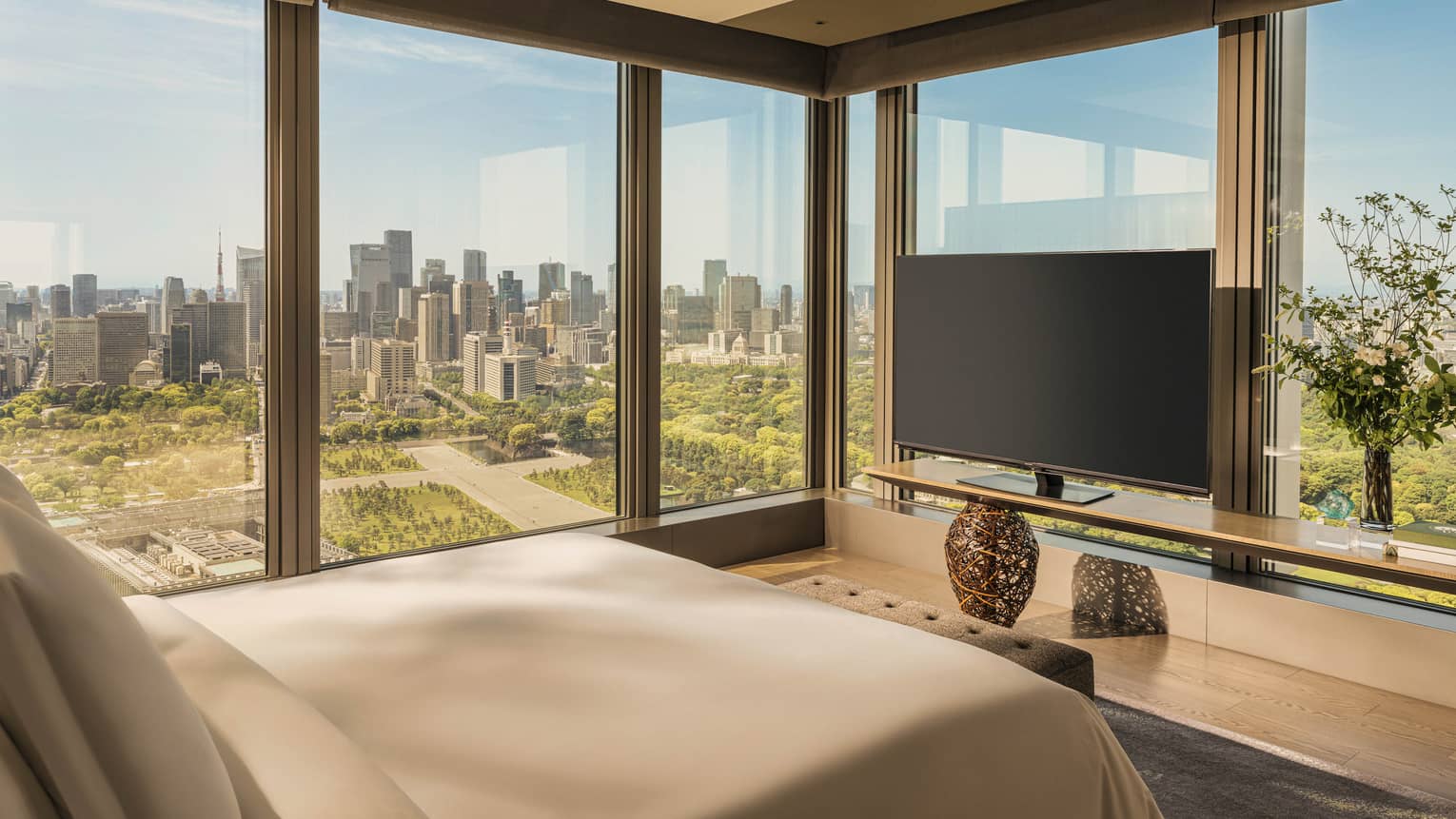 Luxury hotel room featuring king bed, TV and two walls of windows looking out to city and gardens