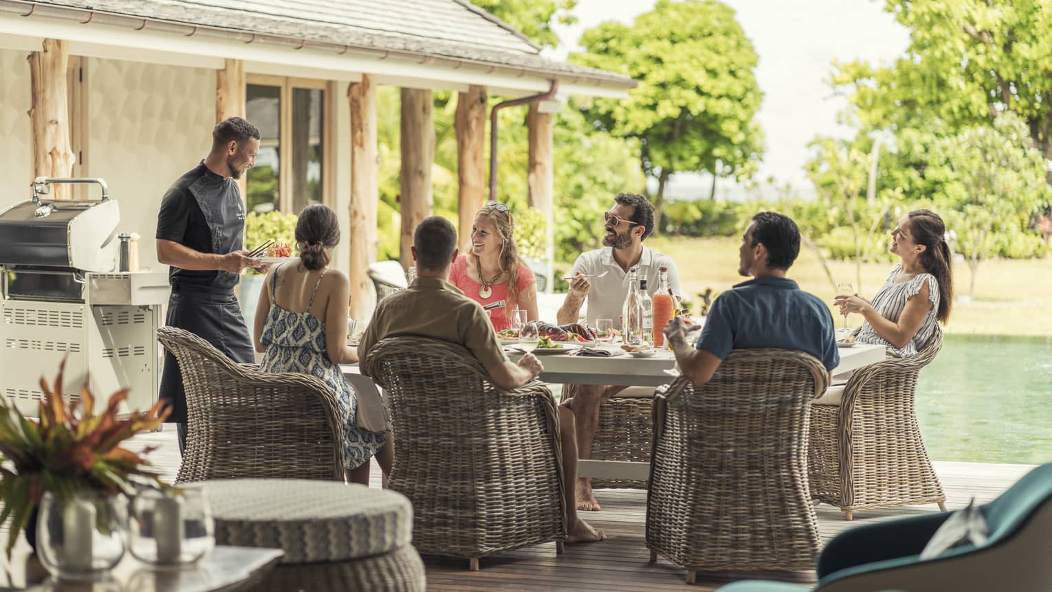 Group of men and women on wicker chairs by pool eating and talking to chef