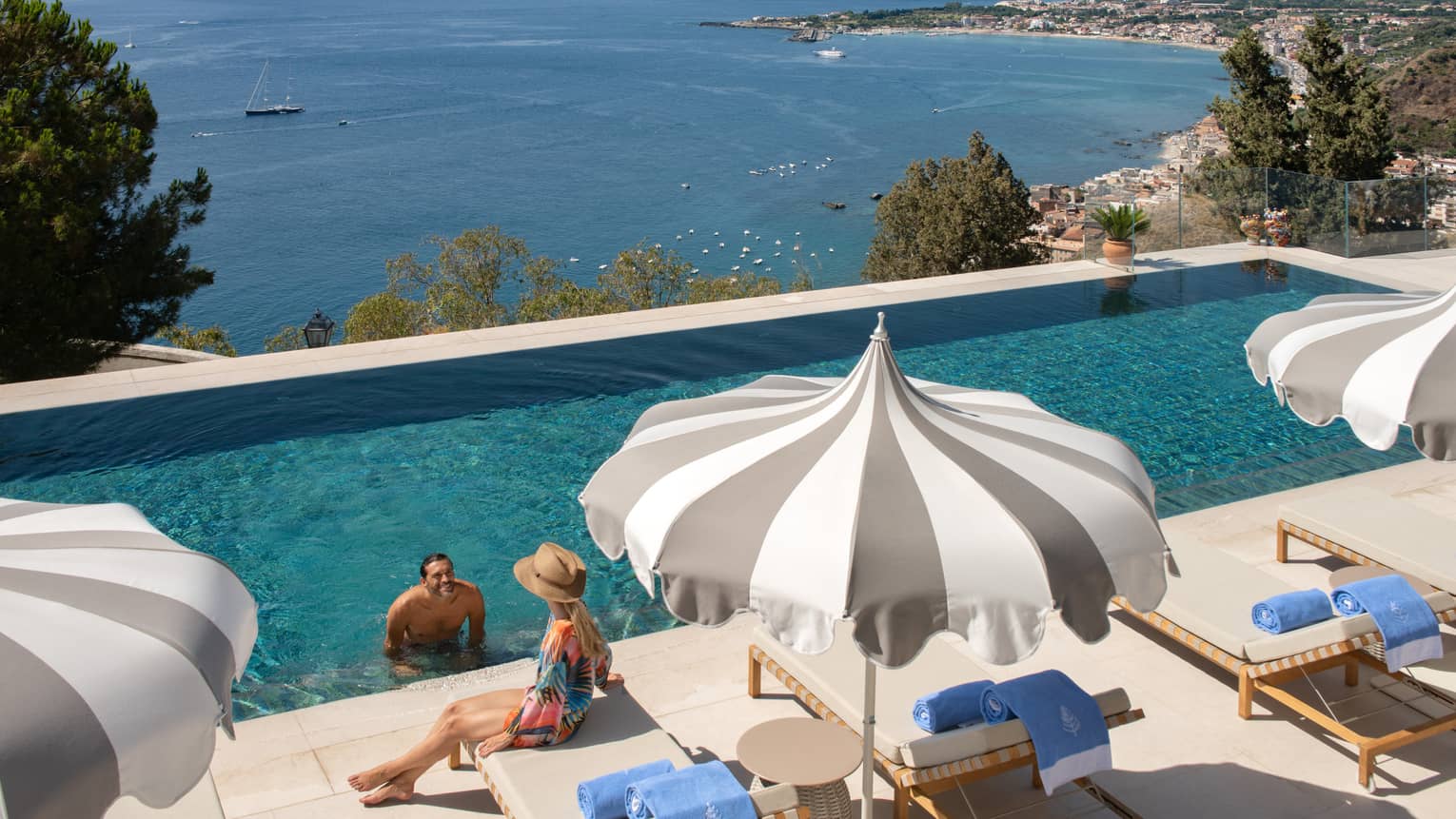A man at the edge of the infinity pool speaking to a woman sitting on a deck chair