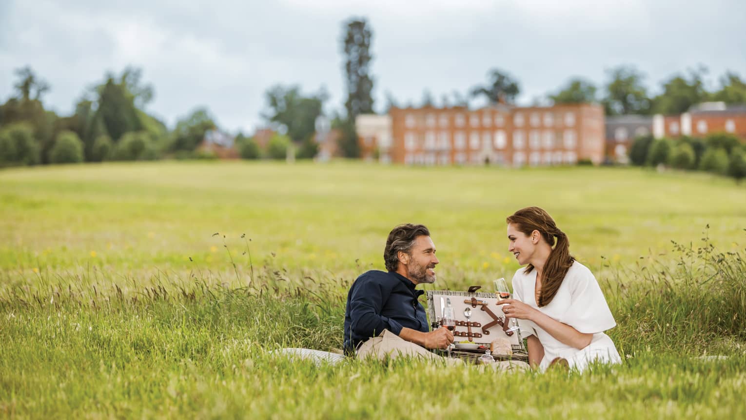 Couple with wine glasses lies in tall green grass with open picnic basket, hotel manor in background