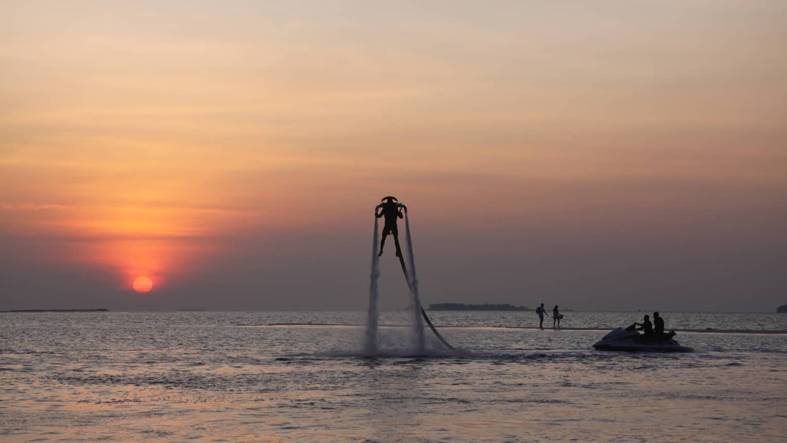 Silhouette of someone with X Jetpack suspended in air over water at sunset