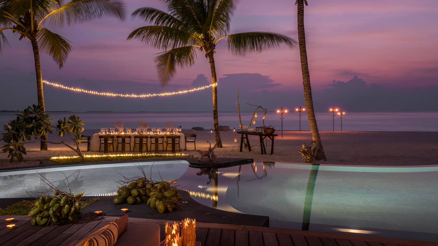 String of lights between palm tree over private dining table on beach by swimming pool at sunset
