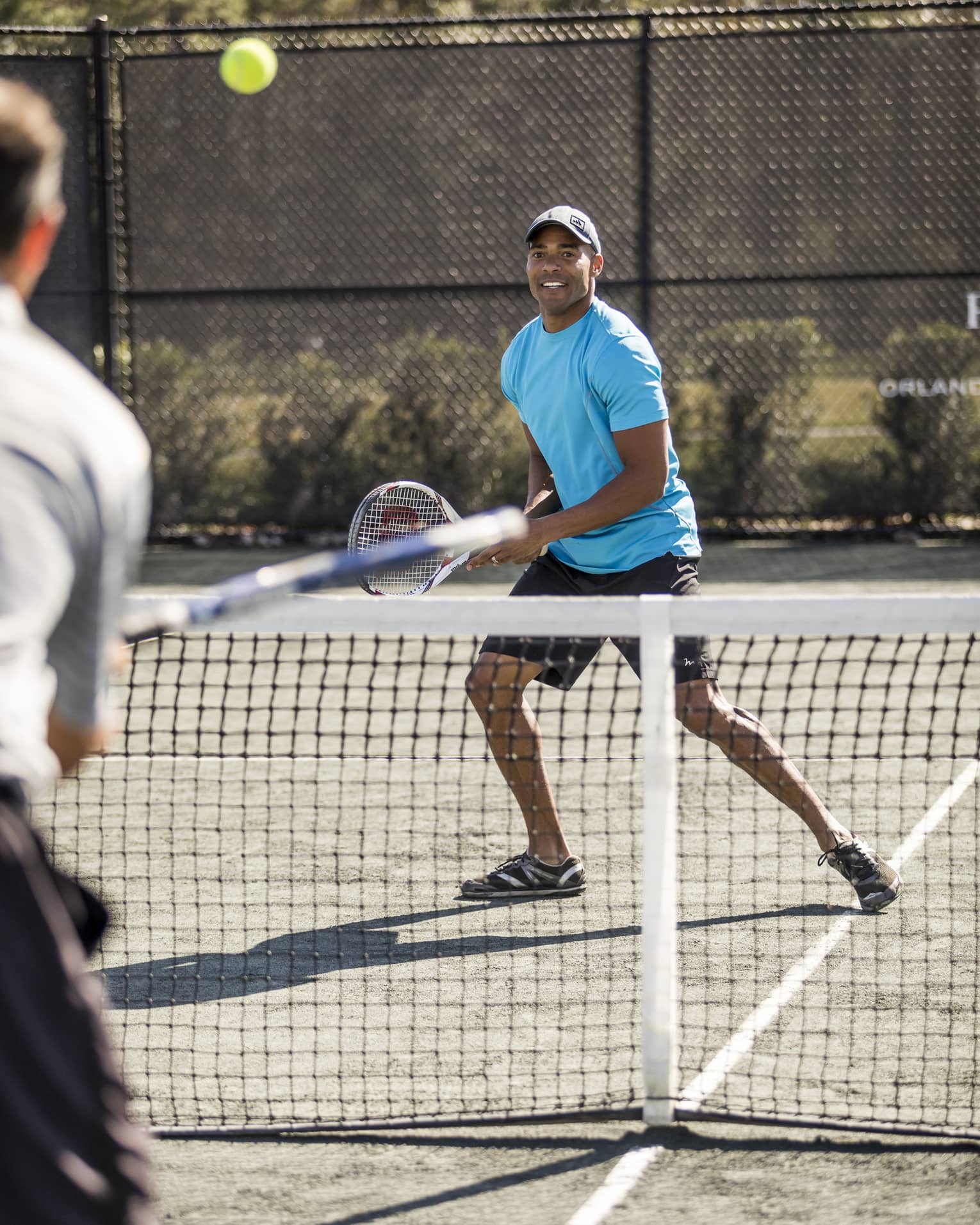 Man wearing blue T-shirt and hat holds racket as tennis ball flies over net