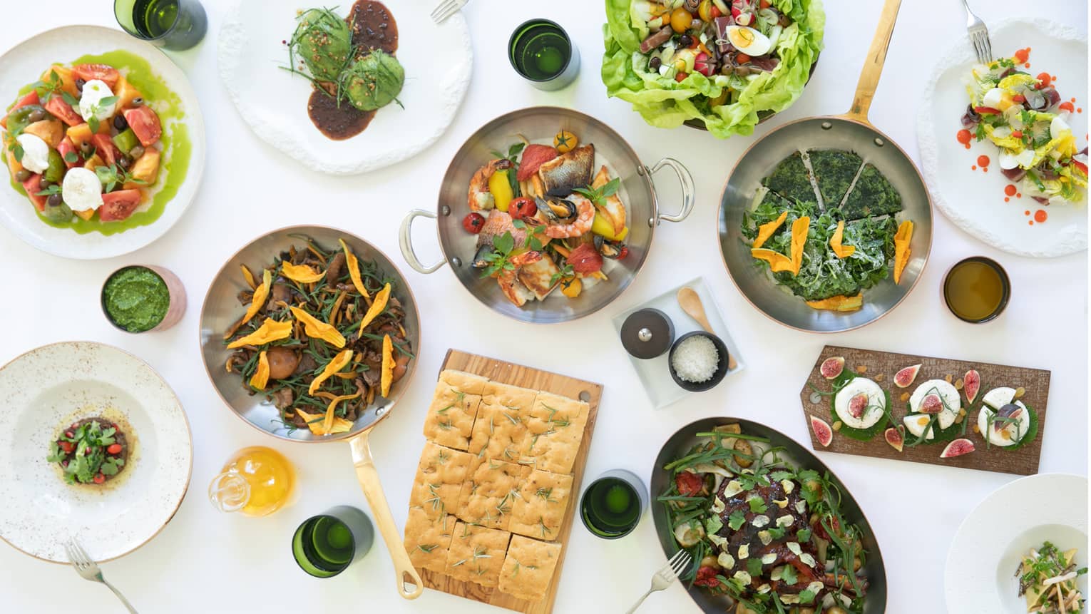 View from above a stark white surface covered in dishes, trays and pans of food, including salads, bread, figs and seafood.