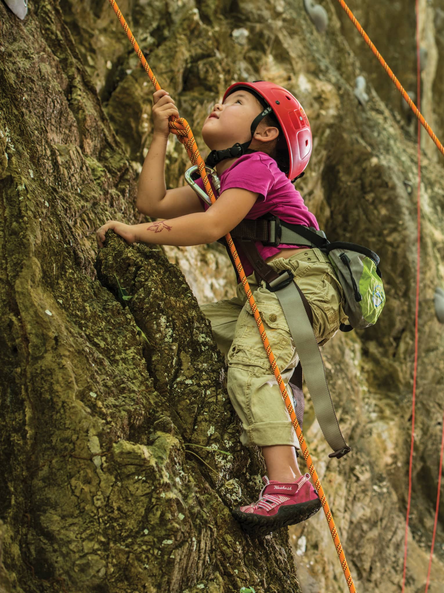 Man holds rope, helps young girl wearing helmet climb rock face
