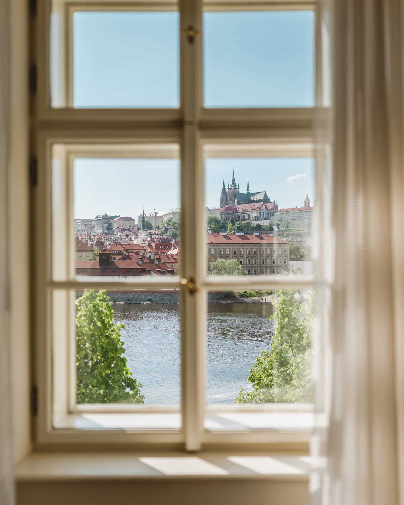 View through a window of a historic city with red rooftops and a cathedral in the distance, overlooking a river and green trees under a clear blue sky.