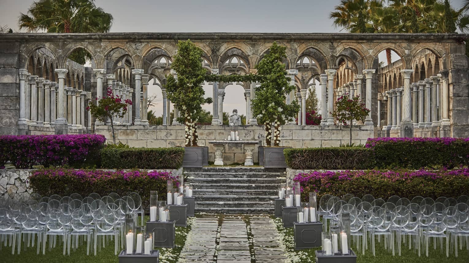Elegant outdoor wedding setup at a historic stone cloister, with rows of chairs, candlelit aisle, lush greenery and blooming purple flowers