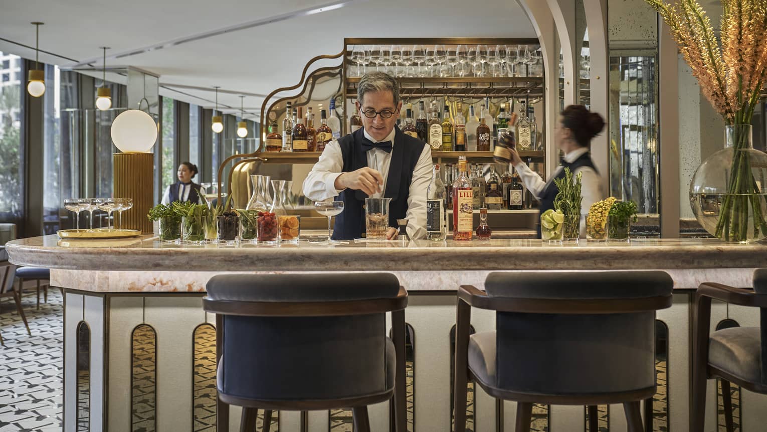 Two bartenders, one male and one female, prepare cocktails at Miss River's marble-topped bar, liquor bottles arranged behind them