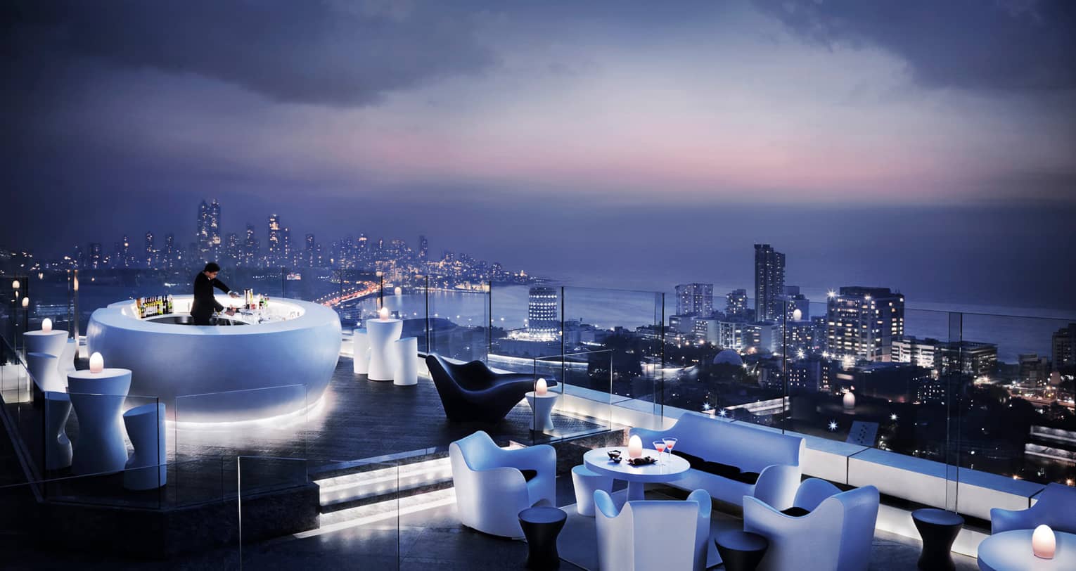 Bartender mixes drink in glowing white circular bar on rooftop lounge, modern chairs and tables