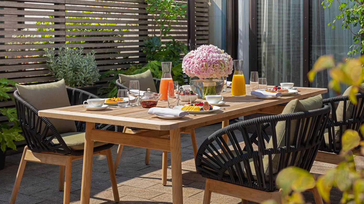 The Executive Conservatory at Four Seasons London Park Lane in Mayfair features an inviting outdoor dining area. A rustic wooden table is set with a colorful breakfast spread, including fresh fruits, pastries, juices, and tea. The table is beautifully decorated with a large bouquet of pink hydrangeas, enhancing the fresh, vibrant setting. Stylish black wicker chairs with comfortable cushions surround the table, nestled amidst lush greenery and a modern wooden slatted privacy screen. The scene is sunlit, offering a pleasant and serene dining experience.