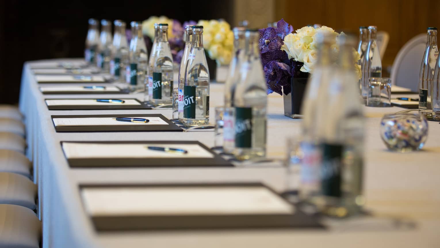 Row of meeting agendas, glass bottles, small vases with flowers on boardroom table