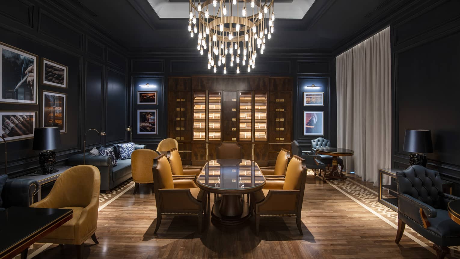 A lounge area with long wood table, dark sofas and walls and a hanging chandelier.