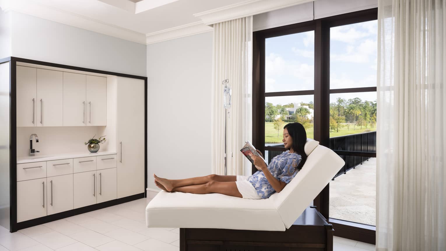 A woman reading a magazine on a folded mattress in a spa clinic with large window.