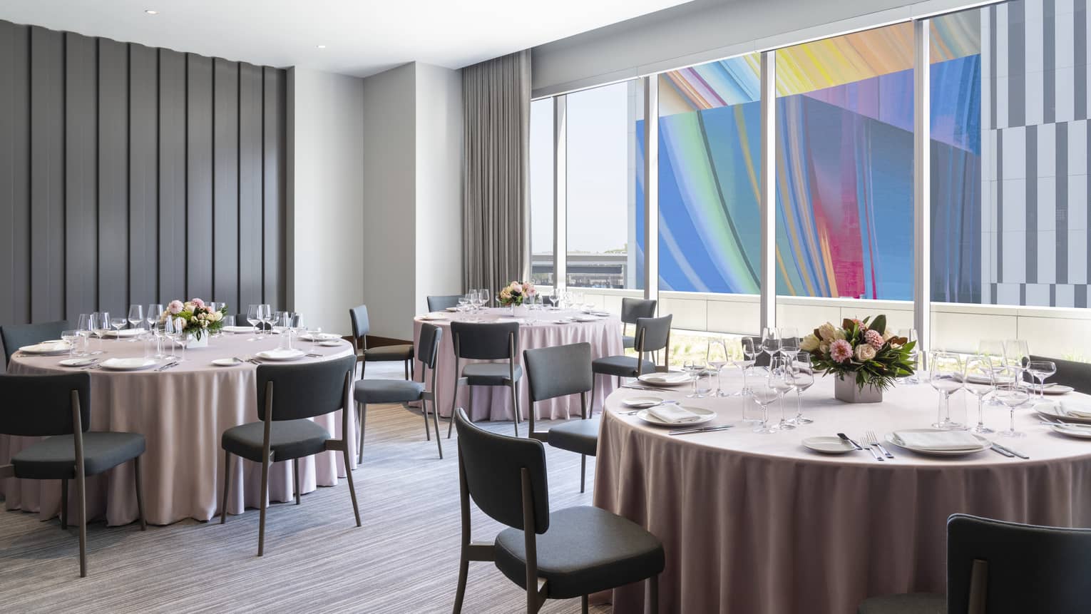 A brightly lit room with round tables and a window facing colourful art outside.