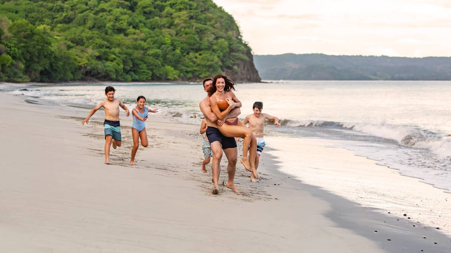 Family of six runs along the waterline on a sandy beach with a tree-covered outcropping in the background