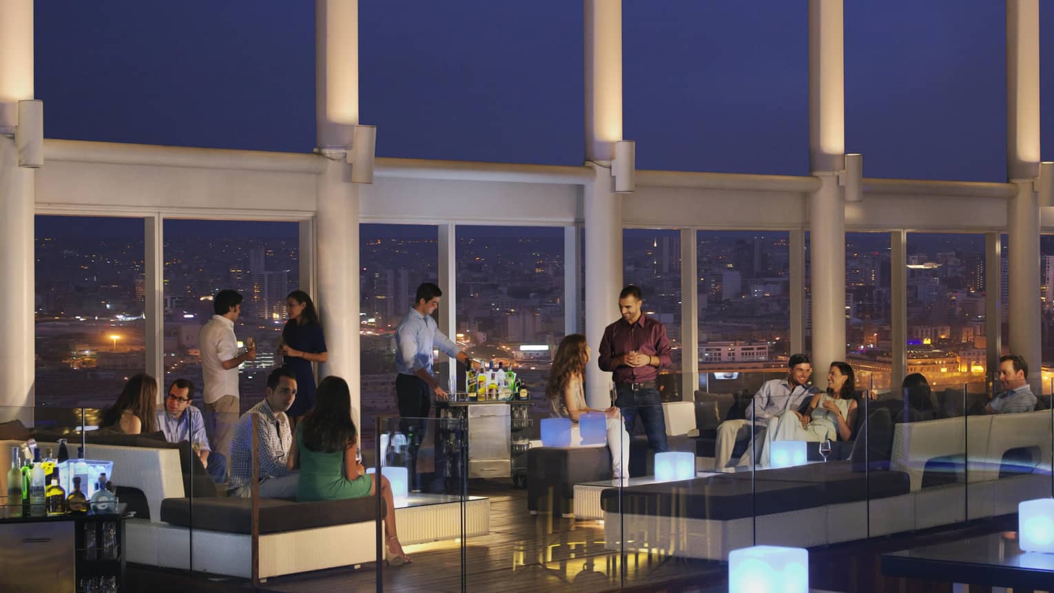 People socialize around cocktail tables, sofas at rooftop bar at night