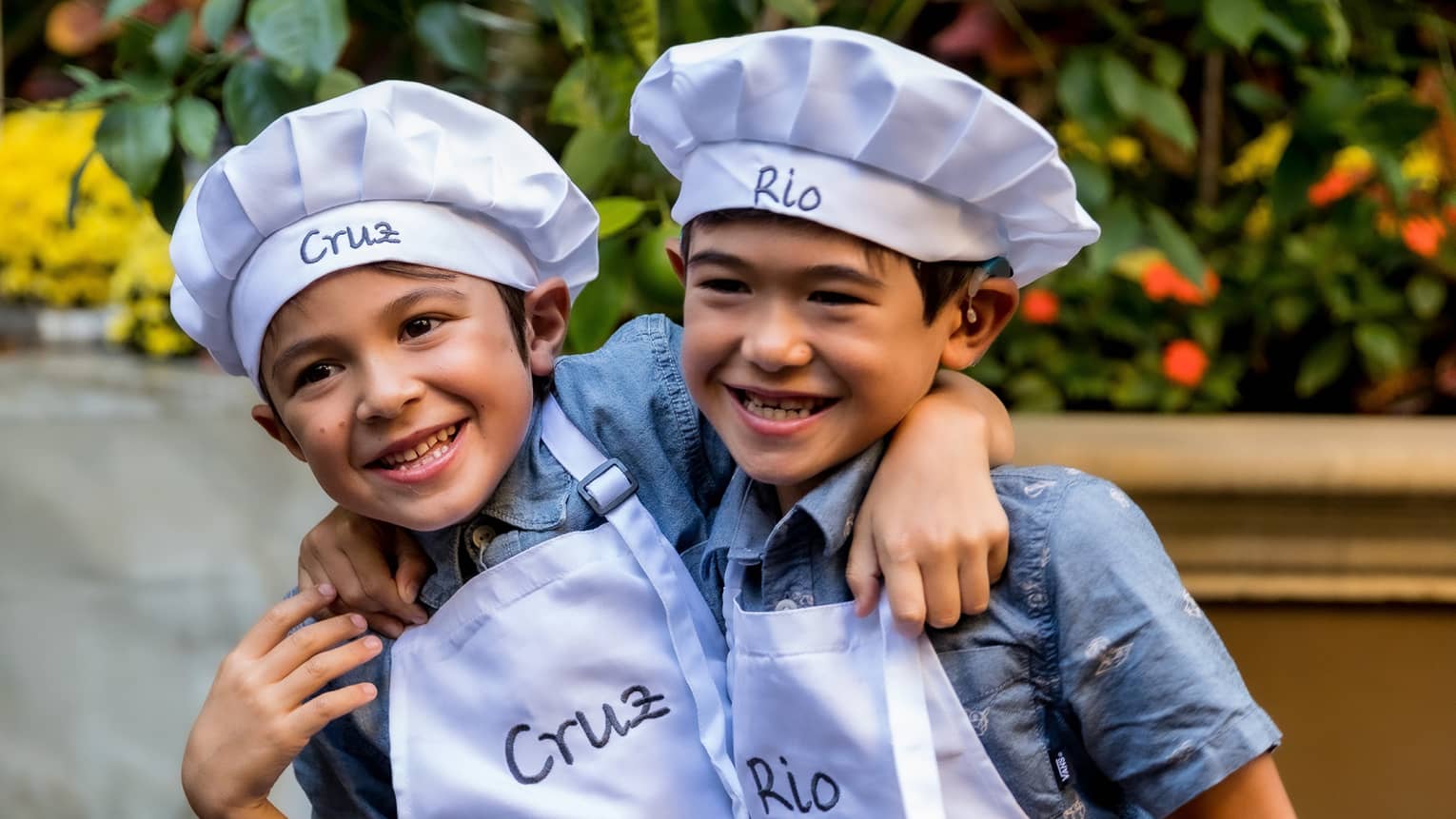 Two children standing together wearing chef?s hats and aprons.