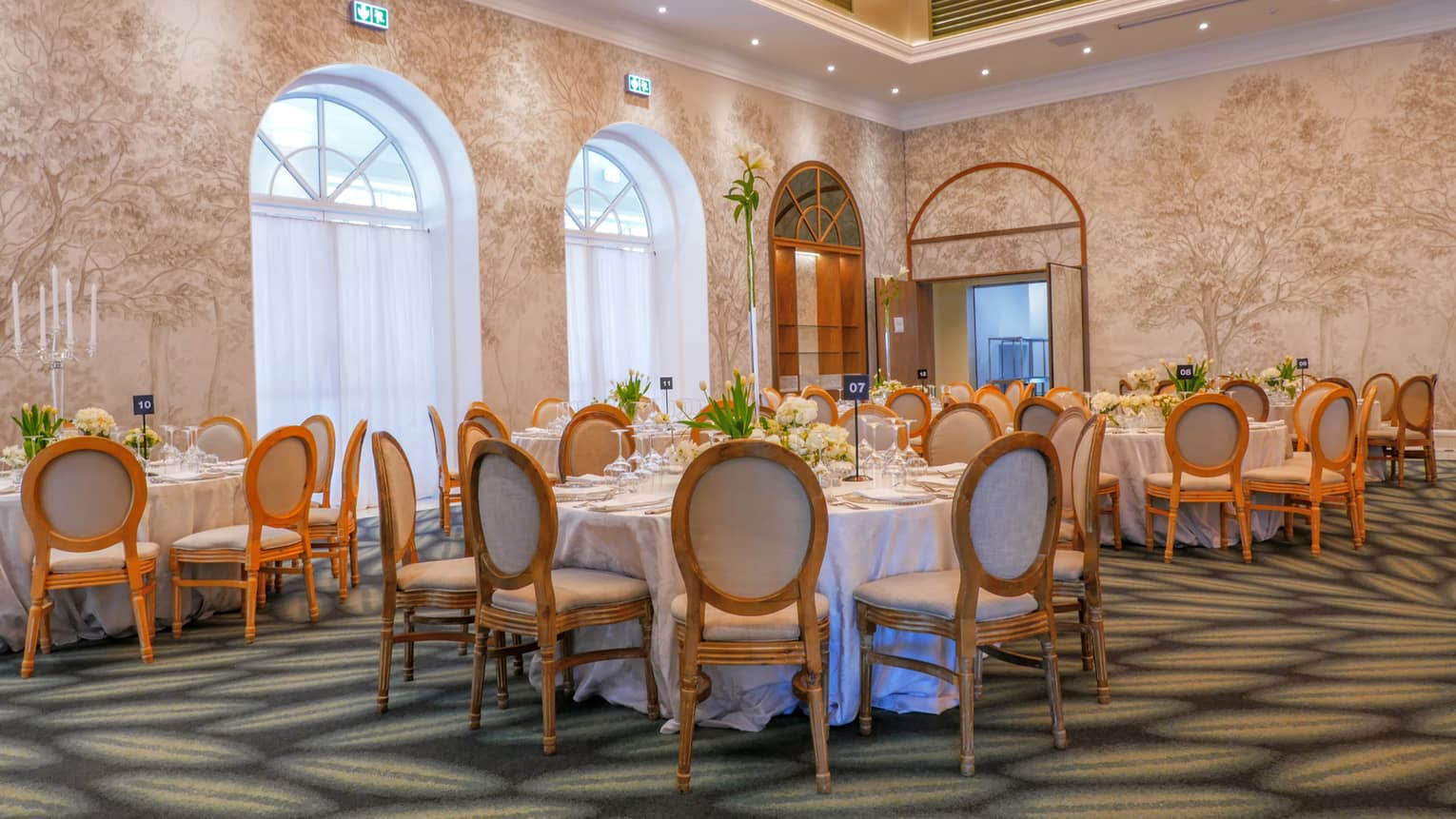 A spacious indoor event room with tall arched windows, patterned walls, round tables with seating, and a patterned carpet