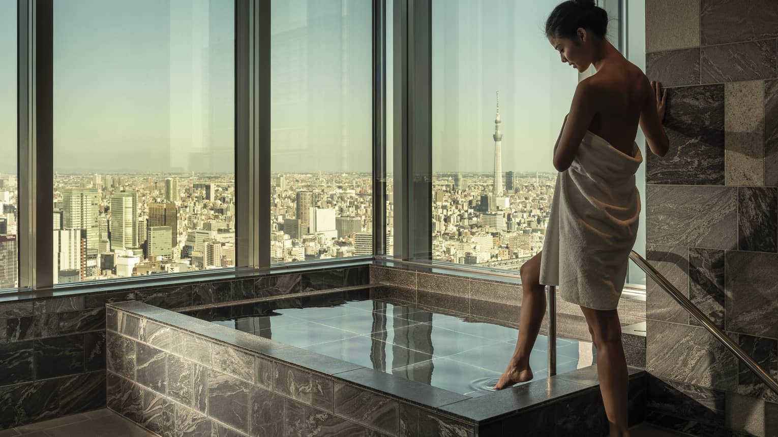 Woman wrapped in white towel dips her toe into the ofuro bath