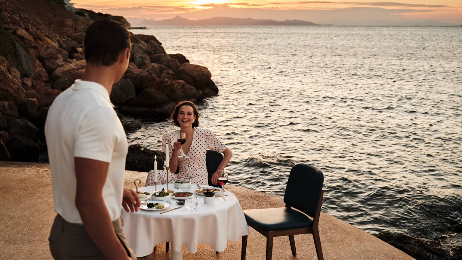 A person sits at an elegant dining table in an outdoor setting, while another person walks towards the table. A rocky shoreline, the setting sun and the ocean are in the background.