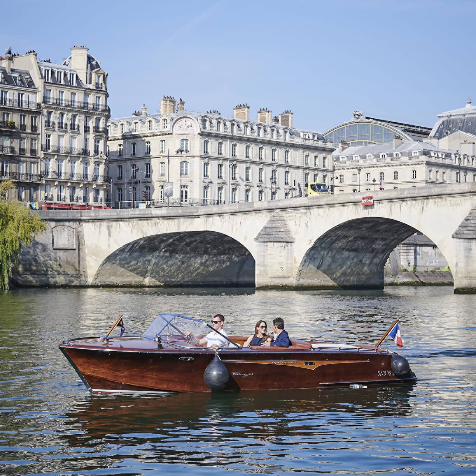 A couple relax in a brown speedboat cruising near a stone bridge along a river lined with white Haussmannian-style buildings.