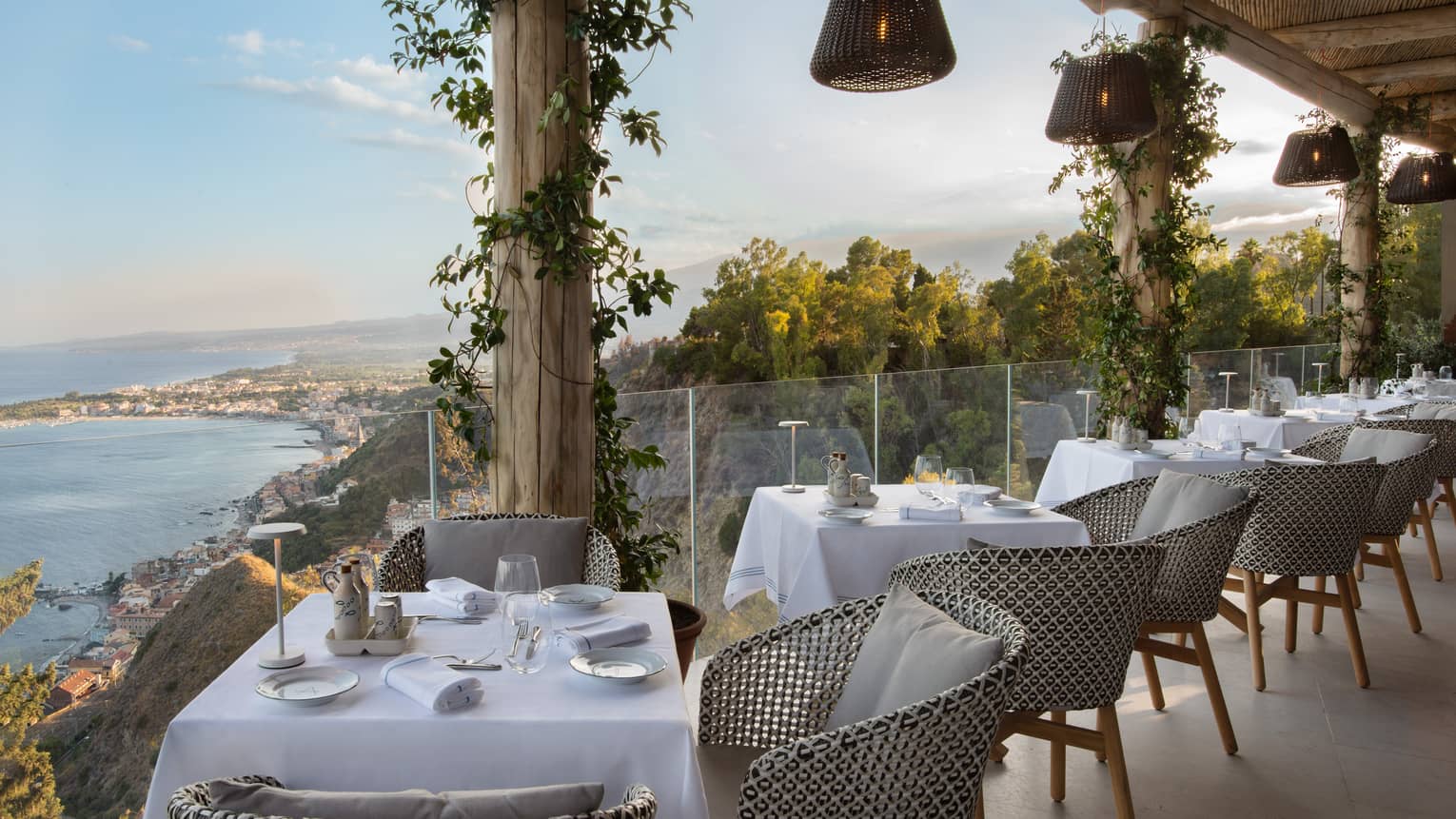 Outdoor restaurant terrace lined with 2-person tables, glass railing and sea view