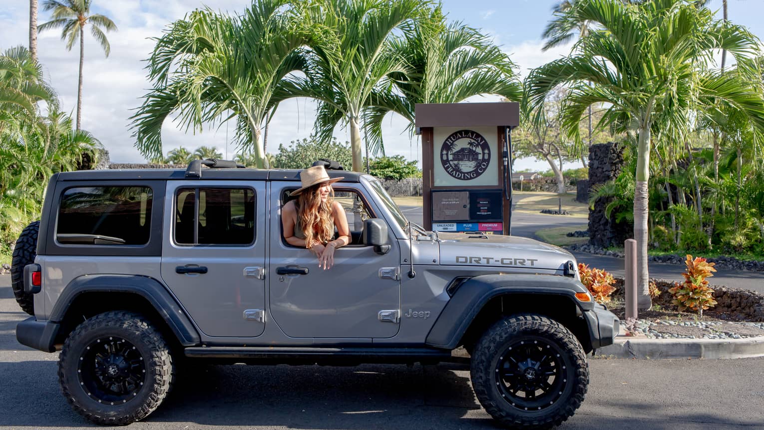 A smiling passenger in a sun hat sticks their head out the window of a silver hardtop Jeep at a gas pump amid palm trees.