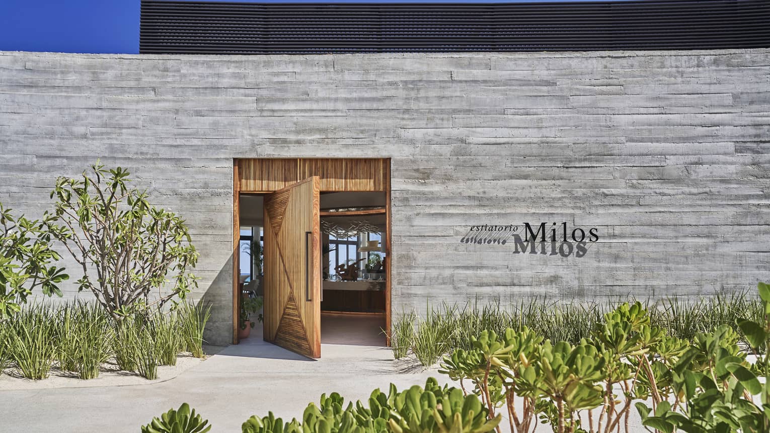 A wood door is open in a grey brick wall, in front of the wall is sand and green plants.