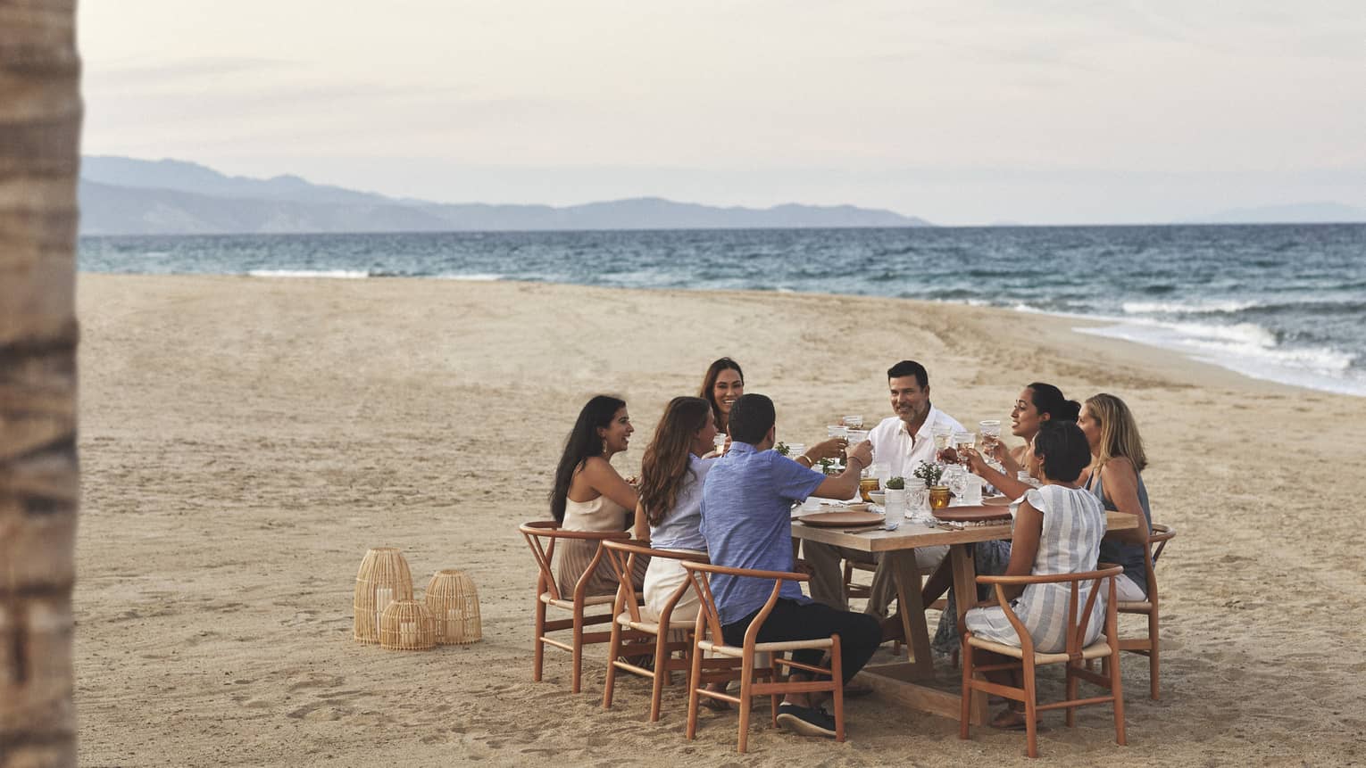 A group dines on the beach in wooden mid century modern dining chairs pulled up around a rectangular wooden dining table