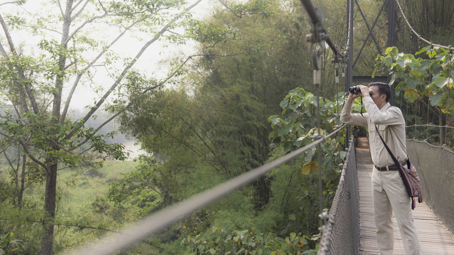 Man wearing artisan bag on suspension bridge surrounded by jungle looking through binoculars 