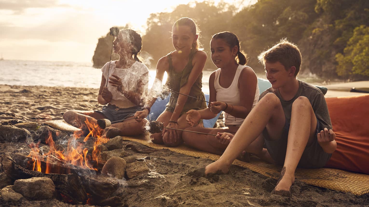 A group of young kids on a beach sitting near a small fire.