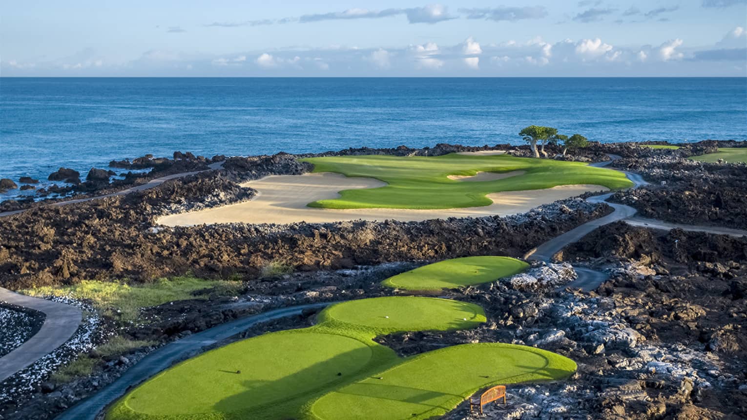 An impressive golf course overlooking the ocean, fairways and sand traps amid black lava formations, blue skies overhead.