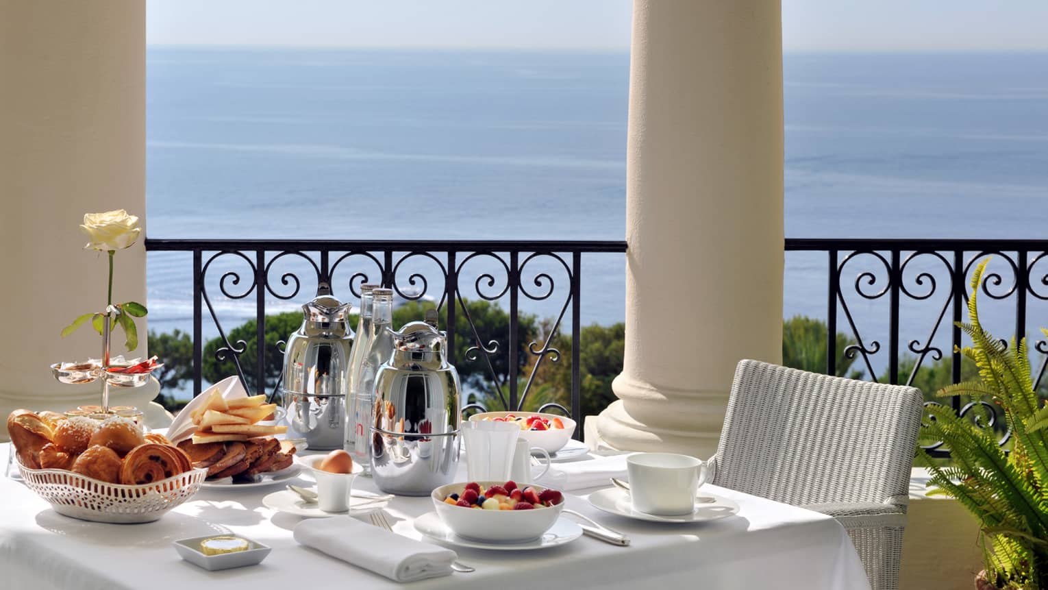 Brunch is arranged on a square table with white table cloth, white whicker chair on balcony overlooking the sea