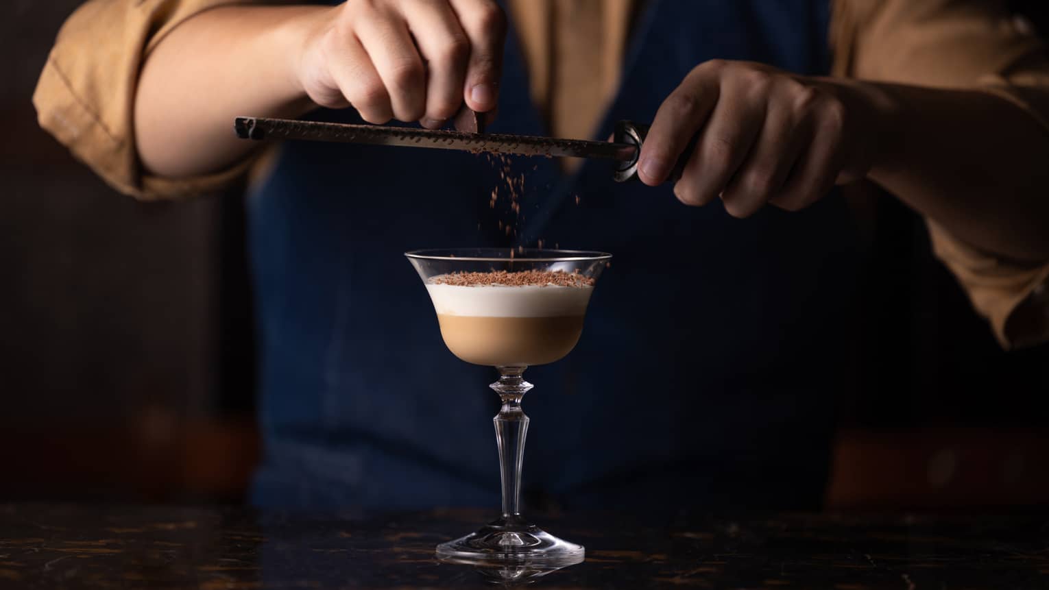 Close-up view of person holding a grater above a cocktail glass and garnishing its two-tiered contents with chocolate shavings