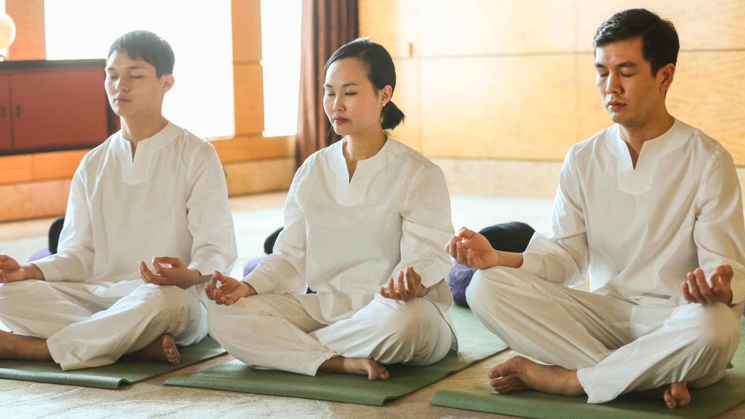 Row of three people wearing white clothing sitting cross-legged, meditating in room