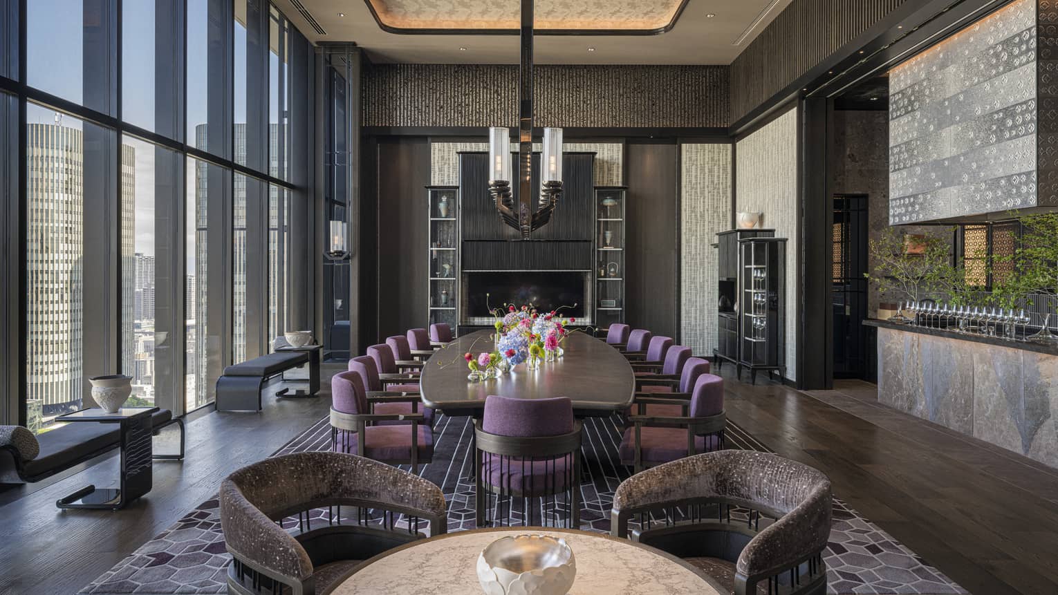A large well-lit room has dark flooring, an area rug, decorative shelving, accents of greenery, and small tables and benches next to floor-to-ceiling windows along one wall, as well as a round table with chairs and a large oblong table with chairs along its length and topped with an assortment of flowers in vases